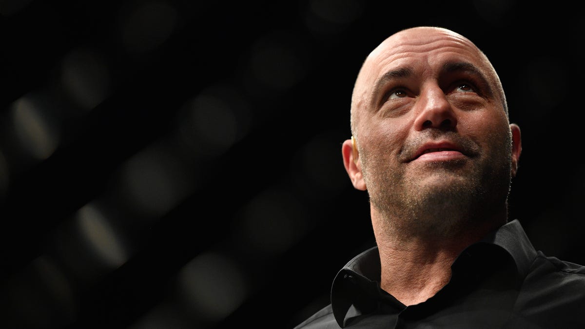 Joe Rogan, in a black button-up shirt, looks up with a smile while standing against a black background.