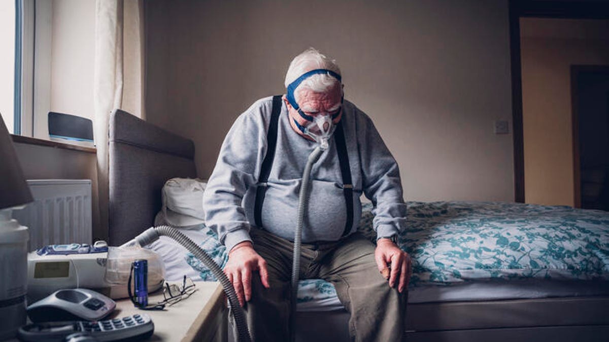 Elderly man sitting on the edge of his bed while wearing a CPAP machine.