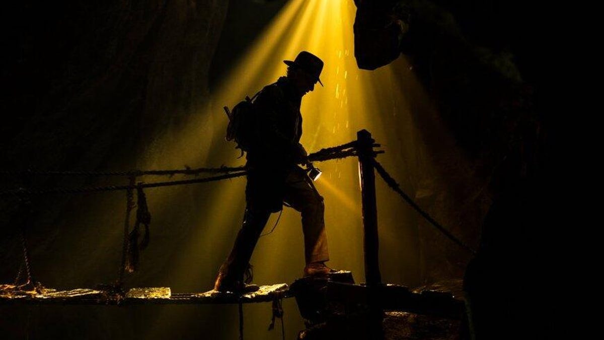 Indiana Jones walks on a rope bridge in a shadowy cave, with a shaft of light hitting him.
