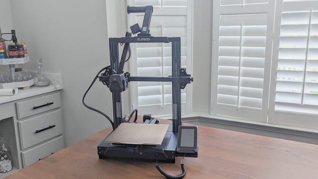 Blue 3d printer on a table with window shutters behind it
