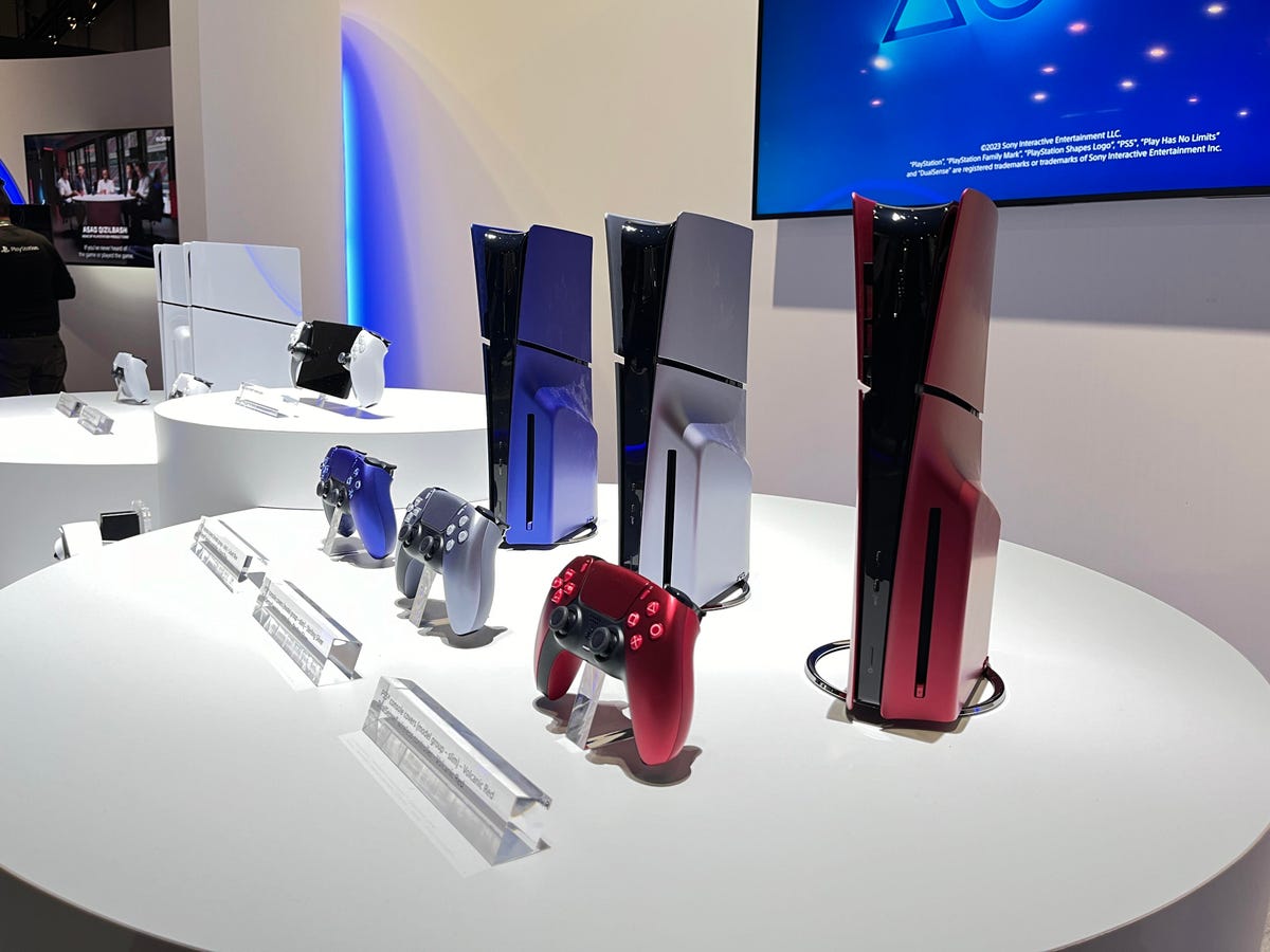Three PlayStation 5s side by side. One is blue, one is silver and the third is red. PlayStation 5 controllers are in front of each