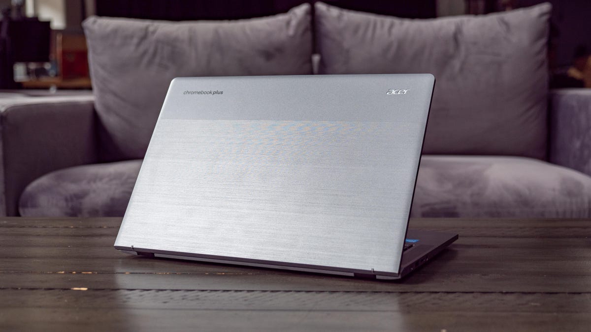 Acer Chromebook Plus 514 open and facing rear on a wood table in front of a light gray sofa.