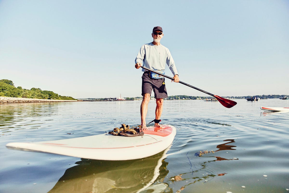 an elderly man stands atop a paddle board in calm water