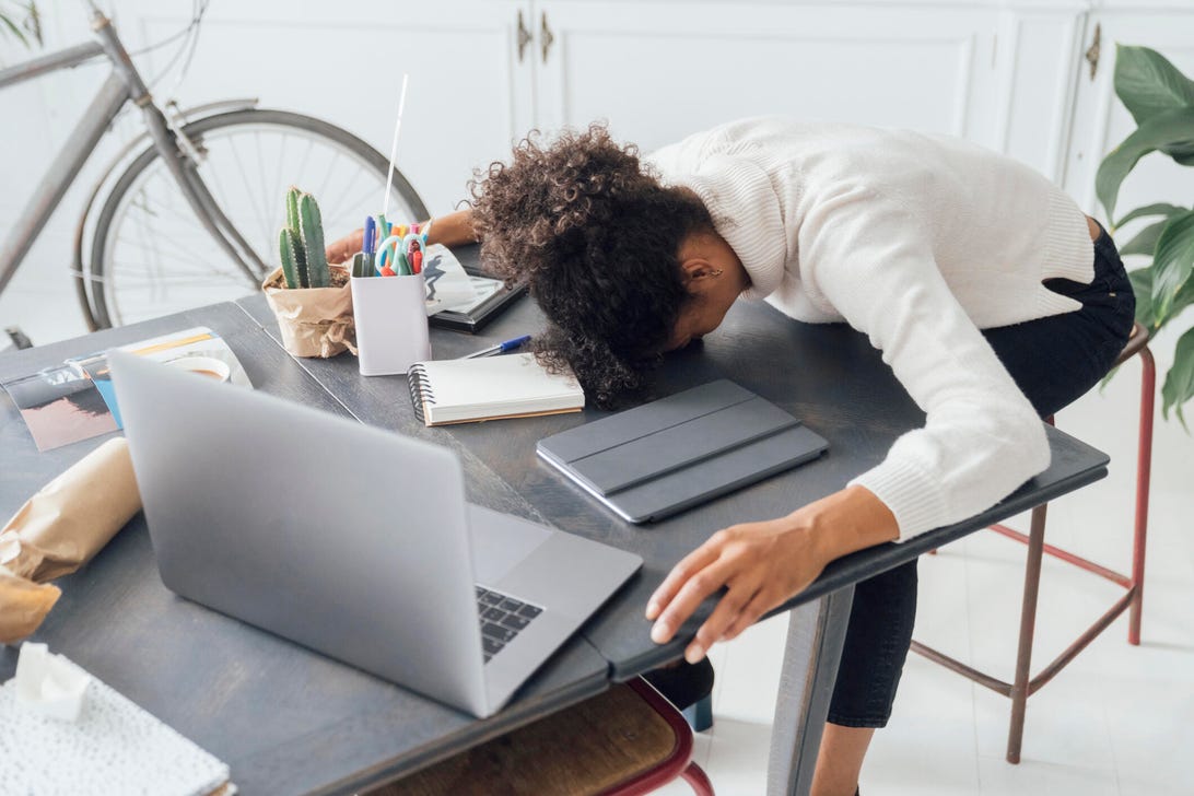 A dark-haired woman in a white shirt slumps over her desk, feeling stressed.