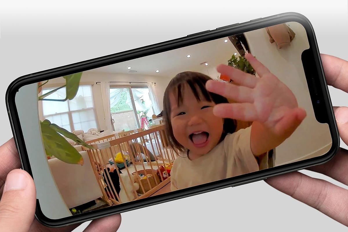 A toddler waves from a living room through the app view of an Arlo home camera.