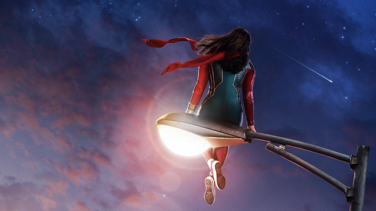 A costumed Ms Marvel sits on a lamp post, gazing at a cloudy sky at sunset
