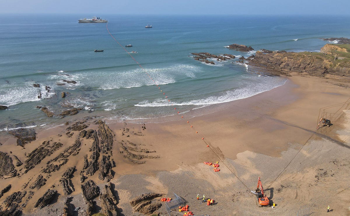 Workers connected SubCom cables supported by orange buoys from a distant cable-laying vessel to a landing point on the beach.