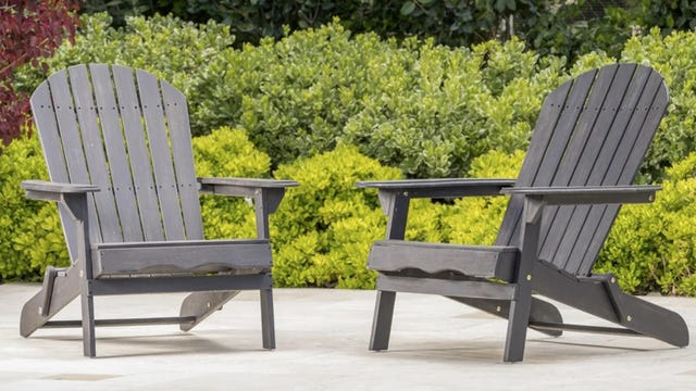 Two gray Adirondack chairs sit on a patio near bushes.