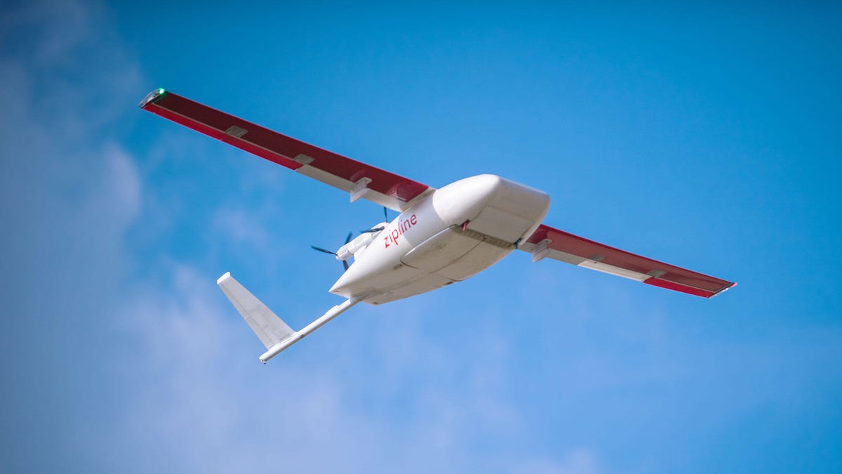 A Zipline delivery drone that looks like a small, red-winged aircraft flies through the sky