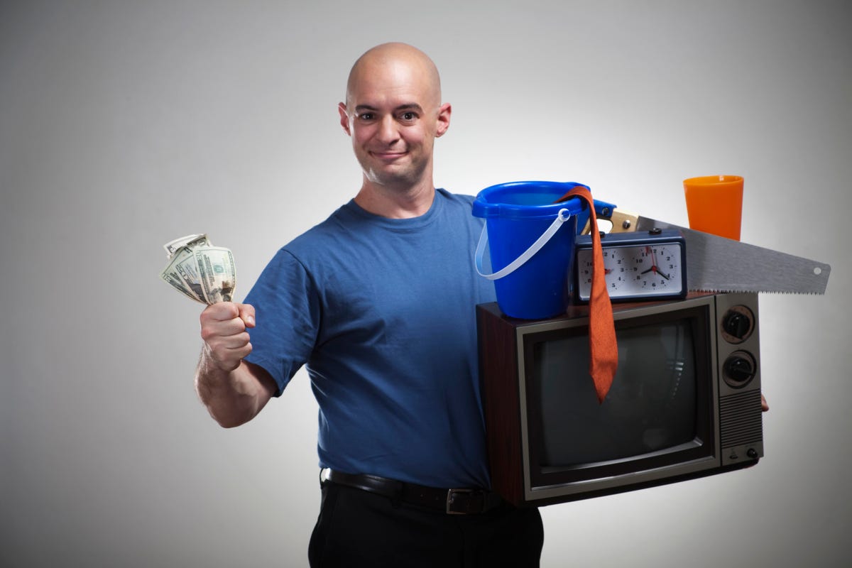 A bald man in a blue shirt holds cash in one hand and a TV with some junk in the other.
