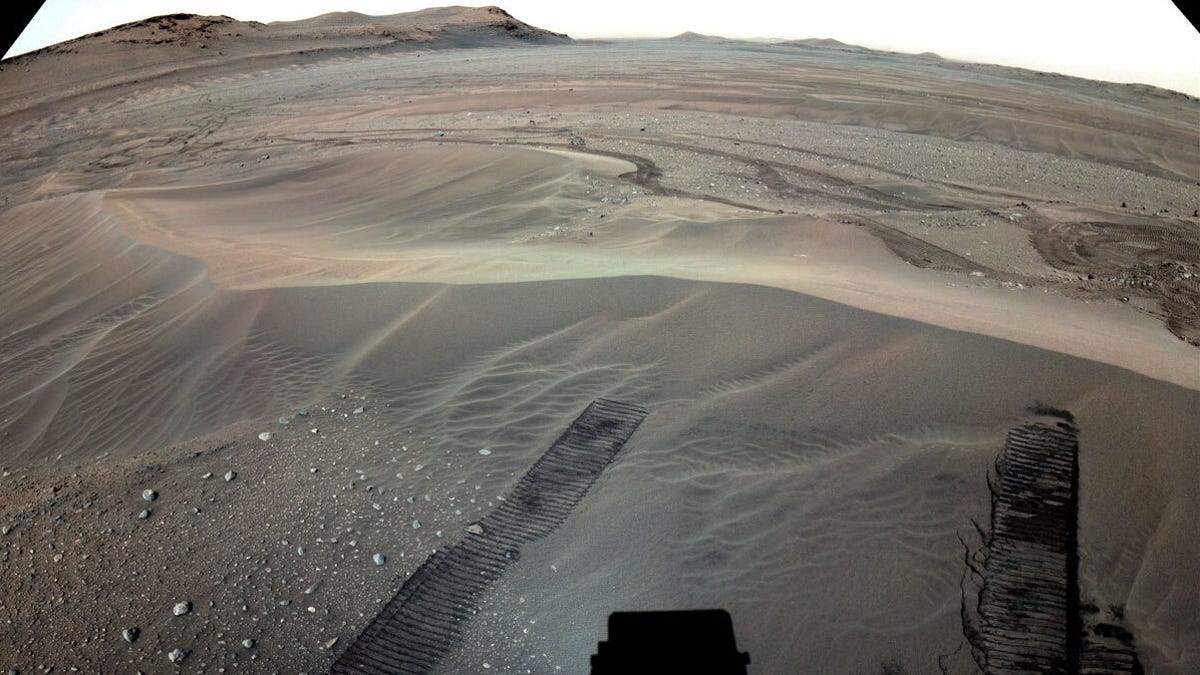 Dark rover tracks cut into a sandy expanse of Mars with distance hills in the background.