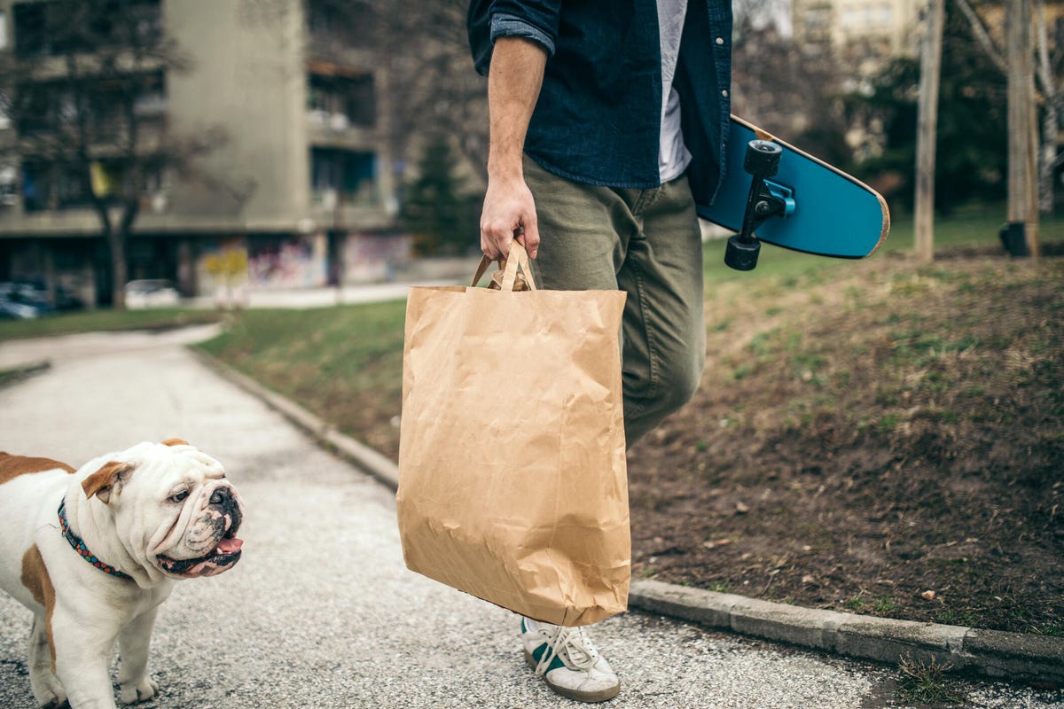 Person walks bulldog while carrying a paper bag and skateboard.