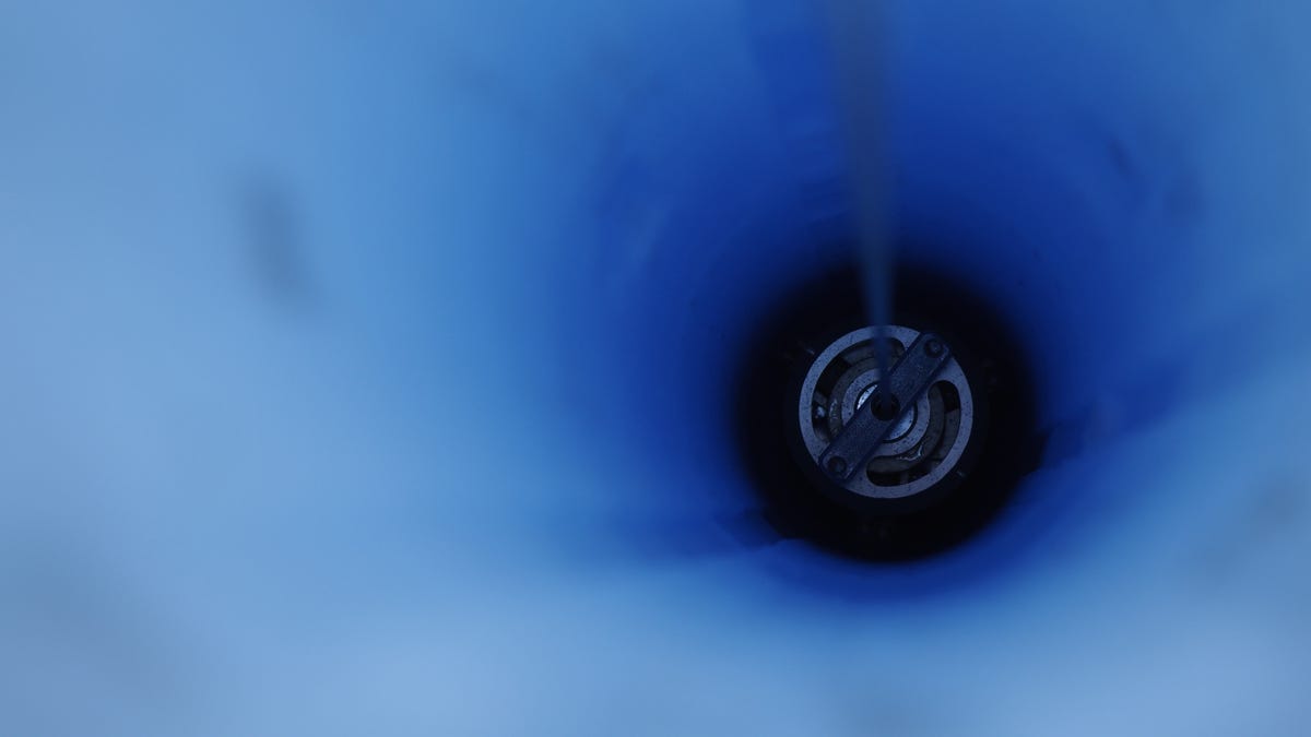 The drill descending into the dark down a borehole which reflects a brilliant, deep blue