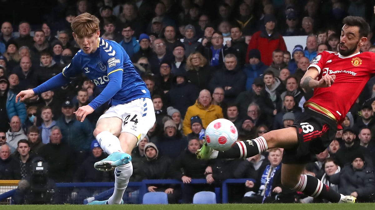 Man Utd's Bruno Fernandes in red and black tries to block a shot by Everton's Anthony Gordon, in blue and white