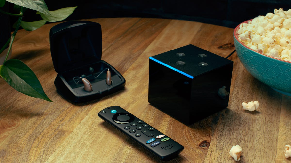 A pair of hearing aids sit in a charging case next to a Fire TV Cube, its remote control, and a bowl of popcorn.