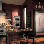 A kitchen with dark wood and red walls features a range, vent hood, double wall ovens, dishwasher and refrigerator from Appliances Connection.