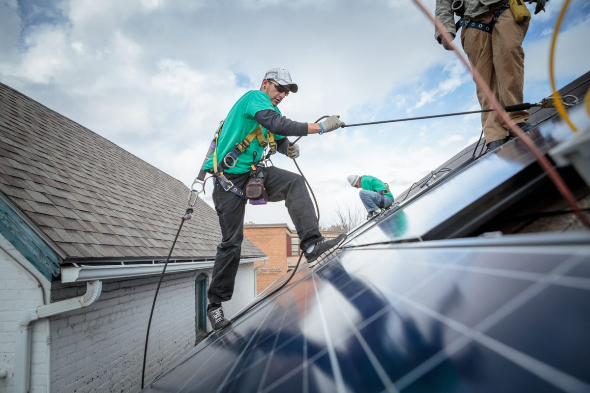 A man in a green shirt climbs a roof with solar panels.