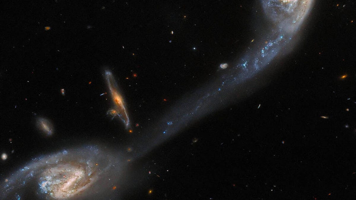 Two galaxies in the foreground appear connected by a wispy, luminous bridge of interstellar dust. A third galaxy is seen in the background, surrounded by many tiny specks of light that represent other, more distant, galaxies across the universe.