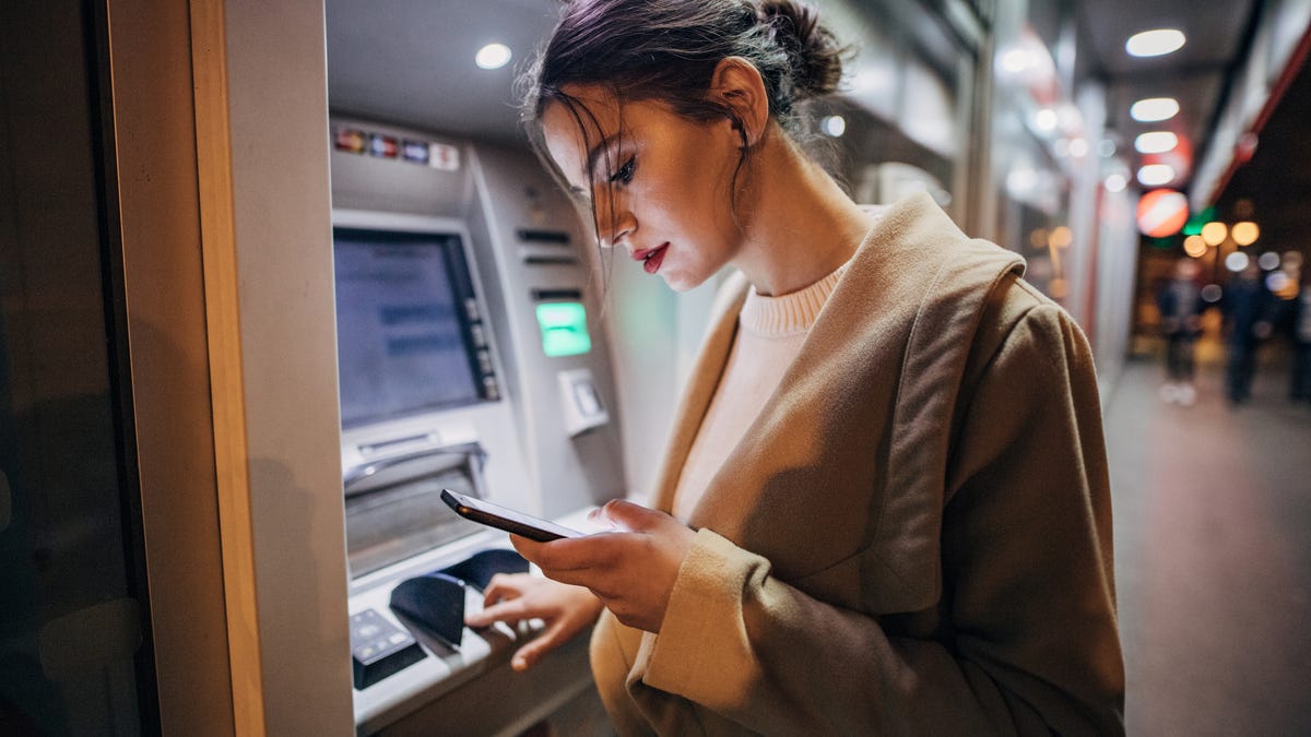 Young woman is using an ATM while looking up something on her cellphone.
