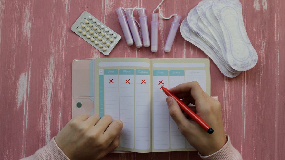 Person uses a calendar to track cycle by hand with birth control and menstrual products nearby
