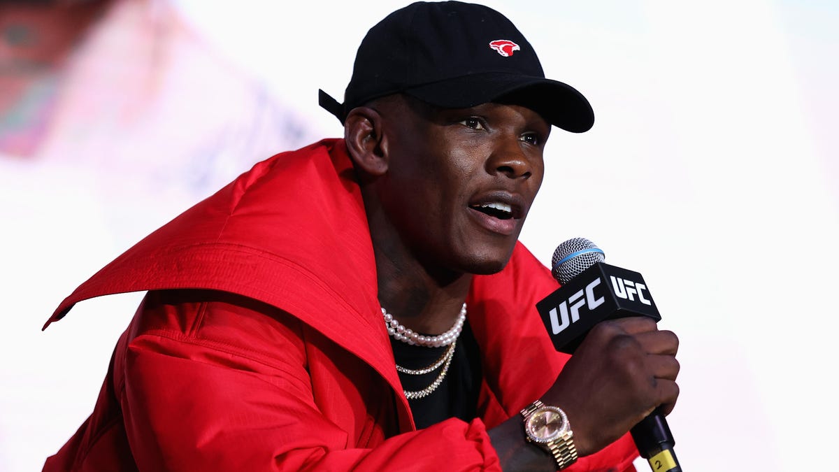 israel adesanya on the microphone wearing a cap and red jacket