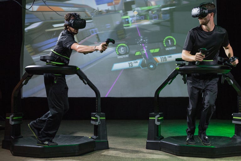Two VR gamers stand in a black treadmill apparatus