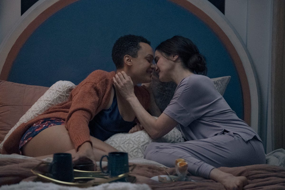 Two women about to kiss in a bed with mugs next to them