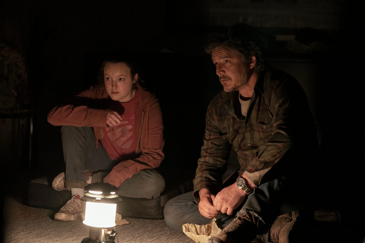 Ellie and Joel are sitting by a lamp in a dark room in The Last of Us
