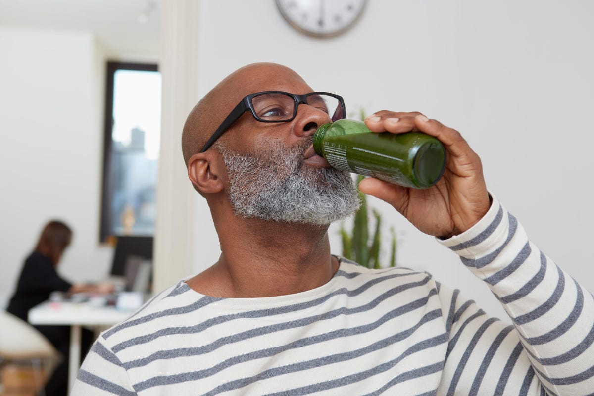 A bald, bearded man in a striped shirt drinks a bottled green smoothie.