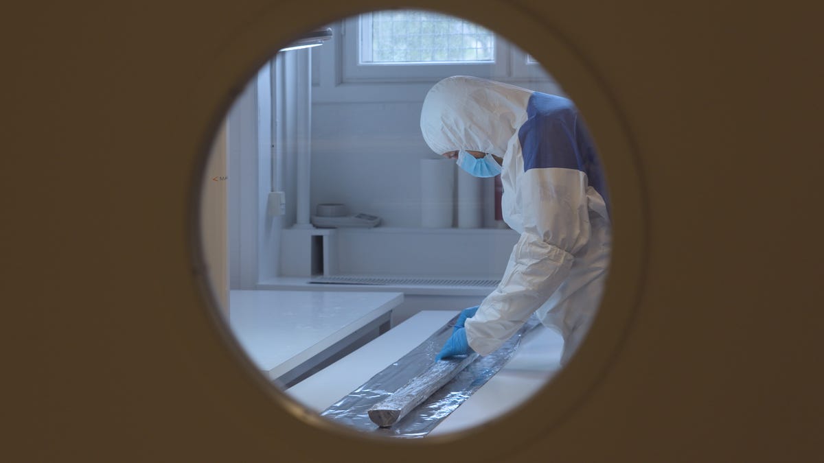 A researcher dressed in a full protective suit, with a blue mask, looks at a sediment core in the middle of a laboratory.  The photo is taken through a circular window.