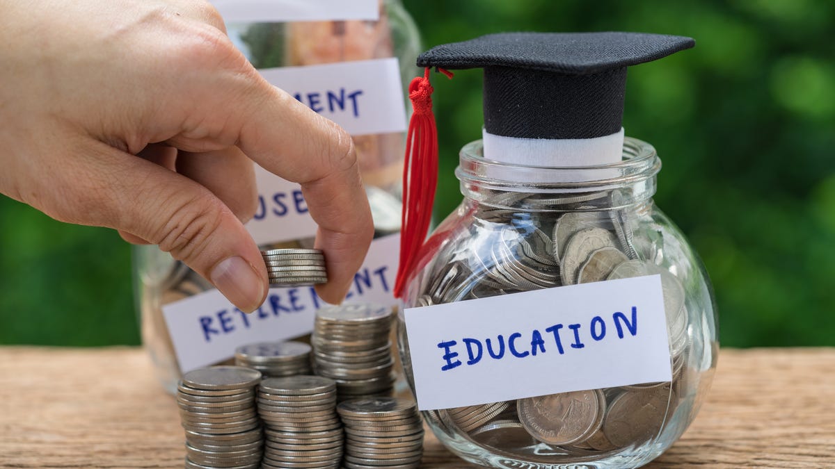 a hand holds a stack of quarters in front of a jar with a graduation cap labeled EDUCATION which sits next to other stack of quarters for saving and retirement