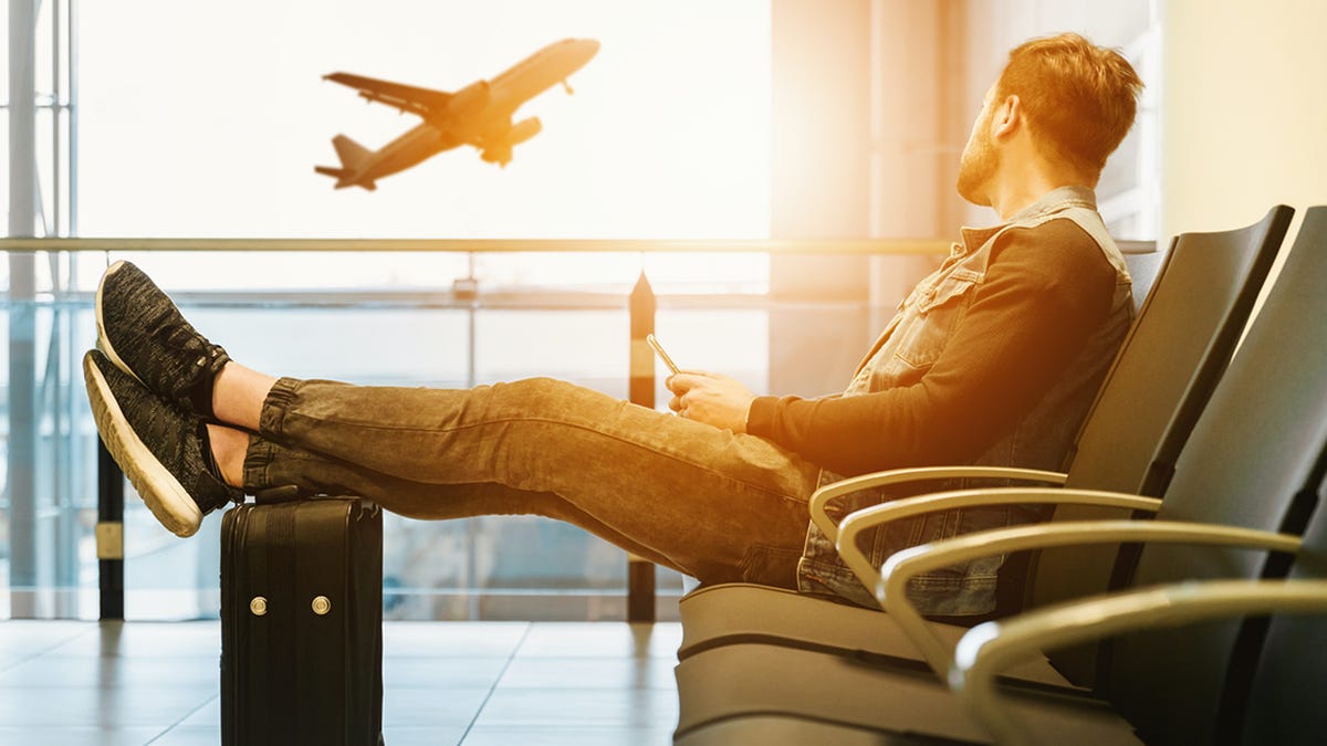 A man with a suitcase sits in an airport and looks out the window, watching a plane take off.