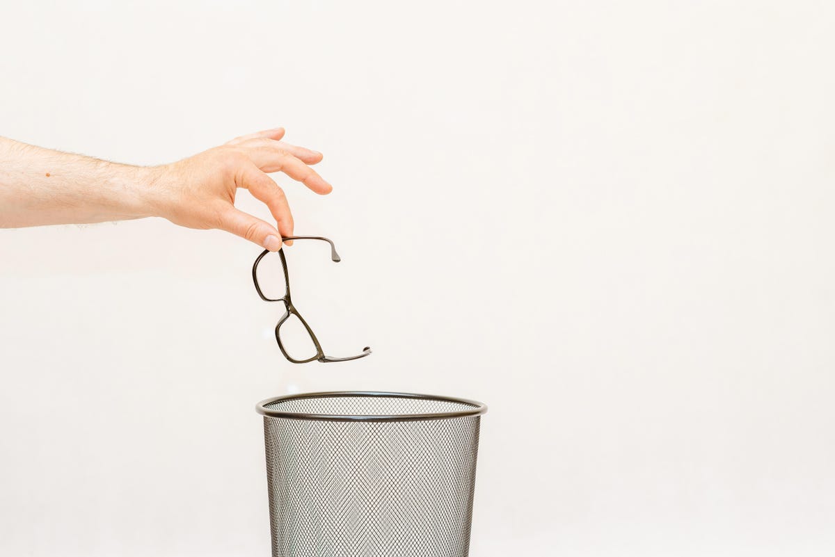 A person dangles their pair of of glasses over a recycling bin