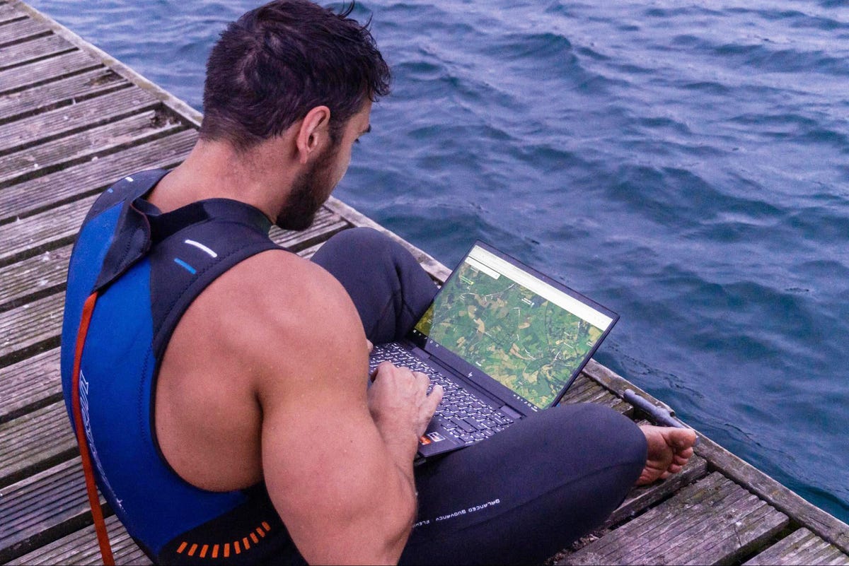 Man sitting on a dock in a wetsuit working on a laptop