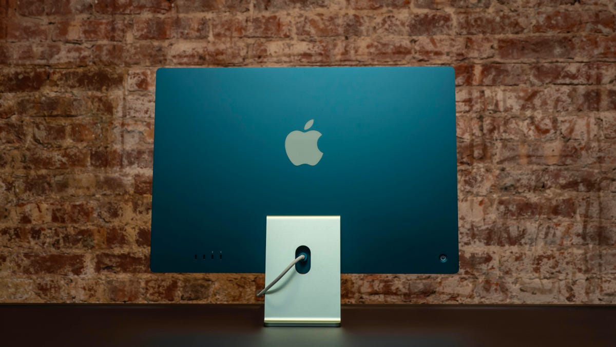 The back of the Apple iMac M3 in green on a black desk in front of a brick wall.