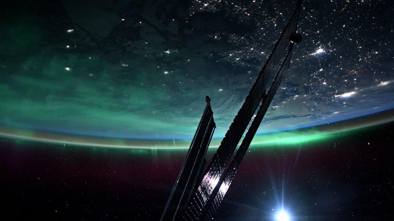 Photo of Earth taken from the ISS shows slim, dark shiny solar panels stretched across dark space lit by a lens flare. Part of Earth is visible upsdide-down with city lights below and green ethereal aurora along the curve.