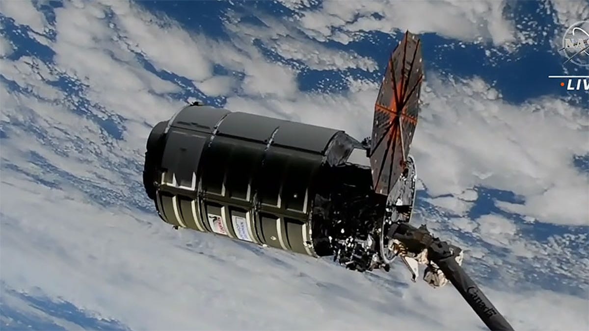 Screenshot shows cylindrical Cygnus spacecraft with one round solar array open and Earth cloudy below.