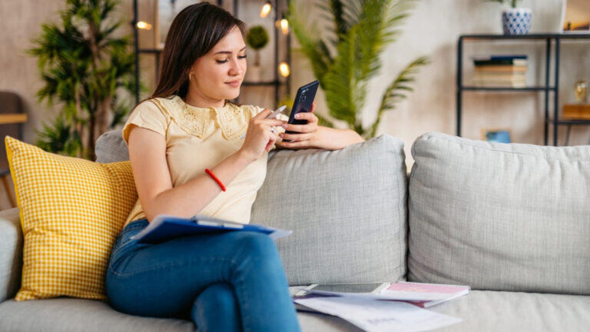 Young woman sitting on the sofa in the living room and filing her taxes on a smart phone.