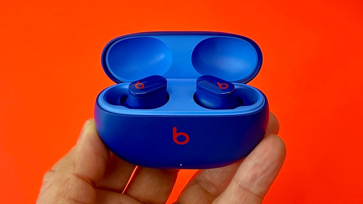 A hand holding up a pair of Beats Studio buds in the charging case against an orange background. 