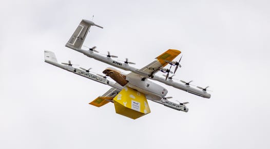 An Alphabet Wing delivery drone carrying a bright yellow package spooled up to rest snugly against the aircraft's underside. Twelve propellers let the drone hover, and four more mounted on the front of its wings let it fly efficiently