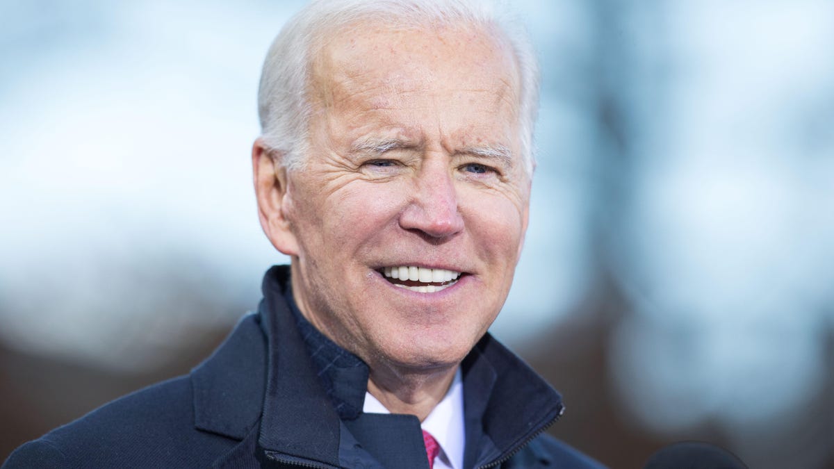 A close-up of US President Joe Biden smiling, with a blurred background of a woodlandlike setting with a blue sky.