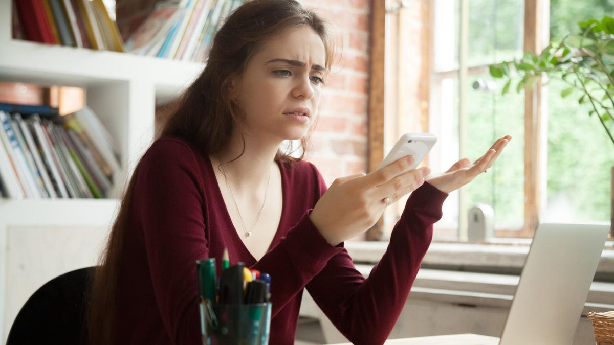 Frustrated young woman looking at her phone while sitting at her home desk where her laptop is open.