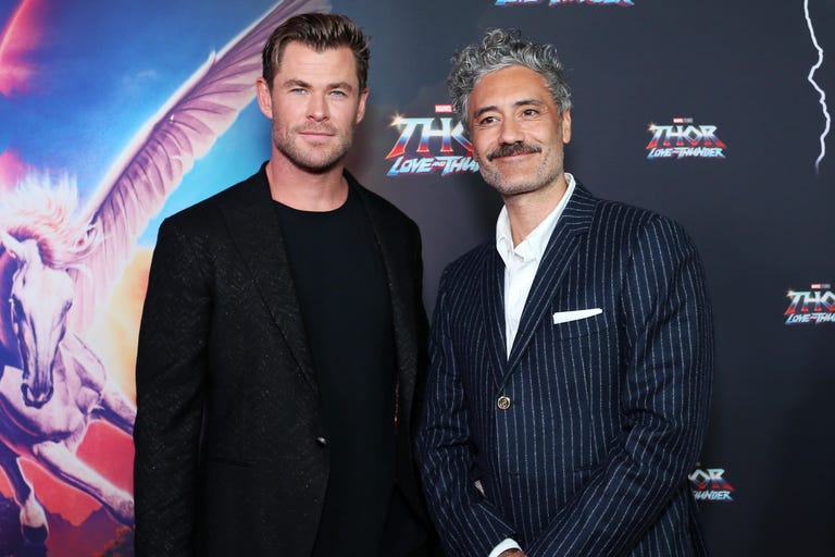 Chris Hemsworth and Taika Waititi in suits standing in front of a poster for Thor: Love and Thunder