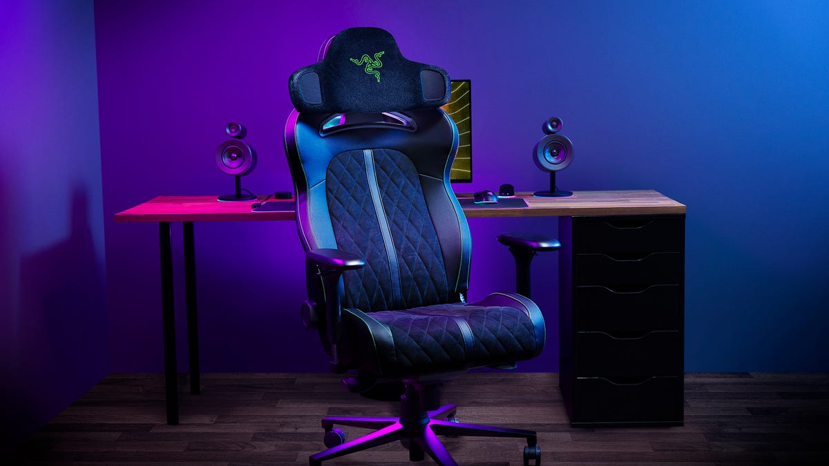 A Razer gaming chair equipped with the Project Carol headrest, facing you, parked in front of a desk under blue and magenta lighting