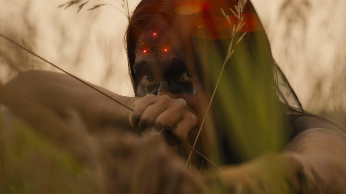 Red dots glare on woman's forehead as she holds a bow and arrow while hiding in reeds