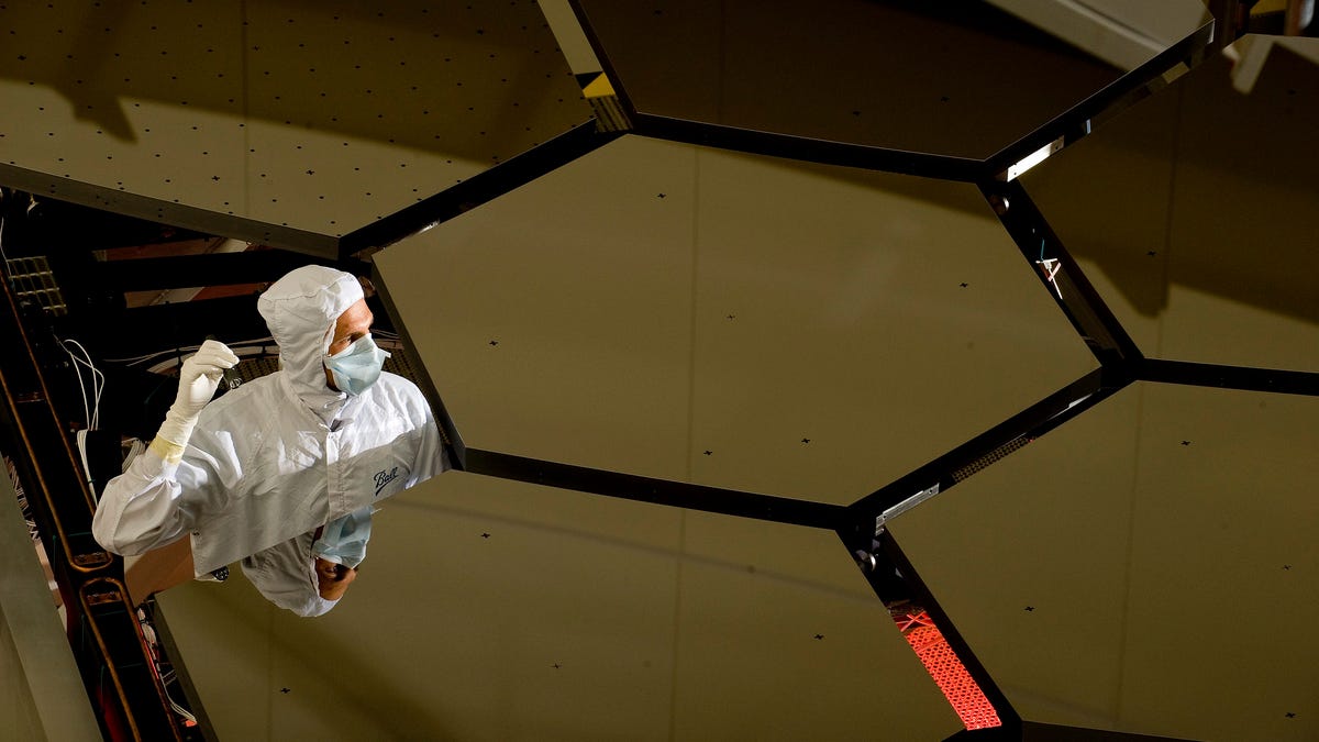 A closeup of six golden, hexagonal mirrors arranged in a honeycomb pattern. On the left side, there's a missing mirror and a scientist wearing a white clean room outfit is standing inside, holding a tool and inspecting the equipment.