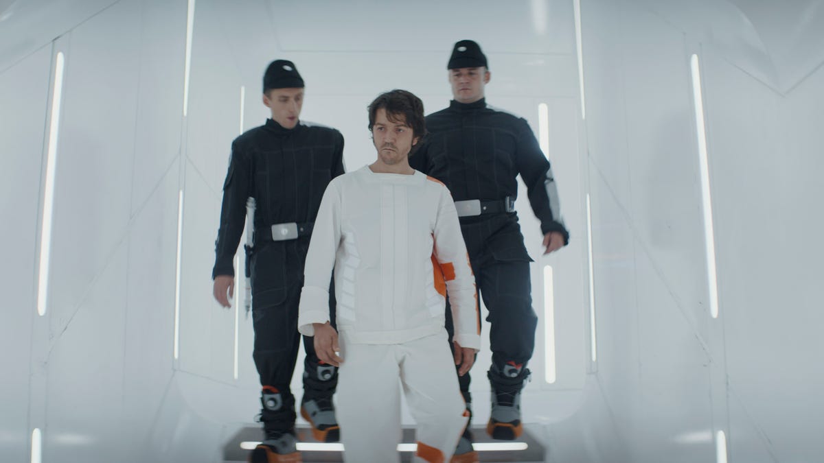 Cassian Andor, wearing a white prisoner outfit, steps into a white-walled Imperial prison while flanked by two black-clad officers in Andor.