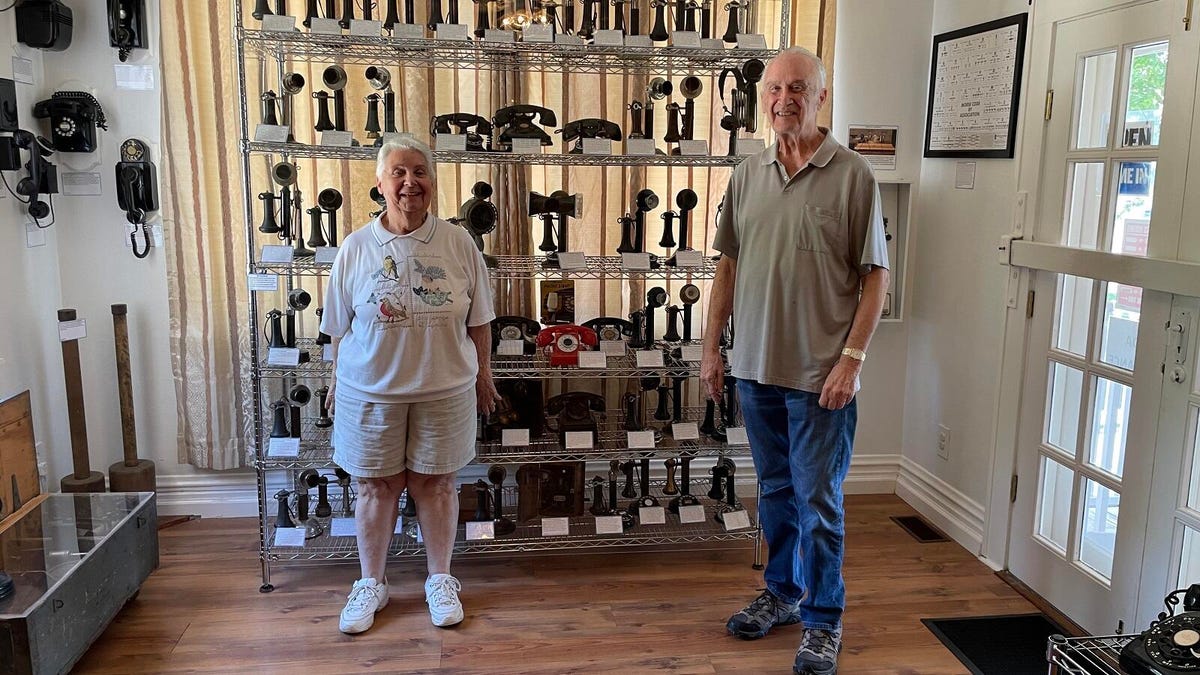 Two people, Carol and Ken, stand in front of old phones from the early 20th century -- models they painstakingly acquired for the museum.