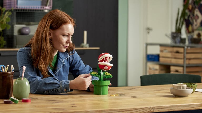 Woman sits at a wood table with a fully built Lego Super Marion Piranha Plant in green and red.