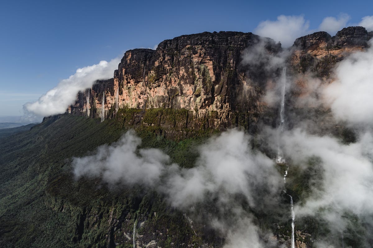 The Weiassipu tepui, a rock mesa on the border of Guyana and Venezuela, is covered in green trees clinging to its walls and waterfalls spilling over it, with foggy mist around.